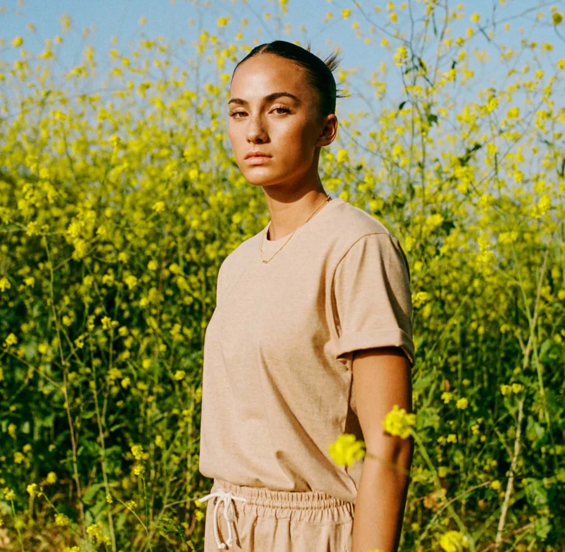 Organic Cotton Beige basic T shirt being worn by a person standing in a field - the brand is Harvest & Mill, a Sustainable Fashion Brand on Sustainable Jungle's brand directory