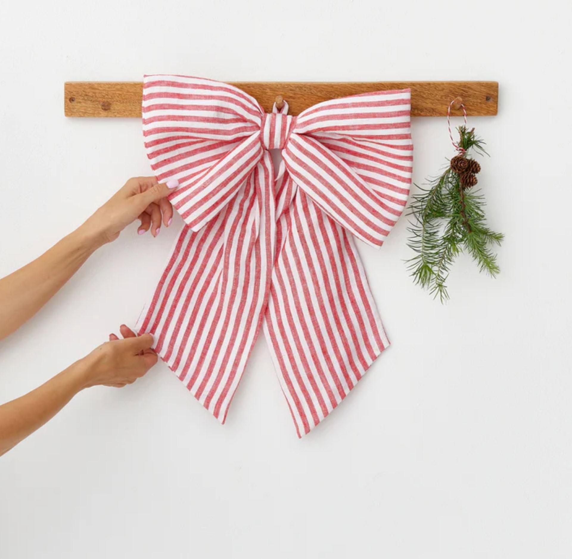 Large red-and-white striped fabric bow hanging on a wooden hook rail with a small sprig of evergreen beside it, showing a simple, reusable, and sustainable Christmas decoration idea. Bow by MagicLinen
