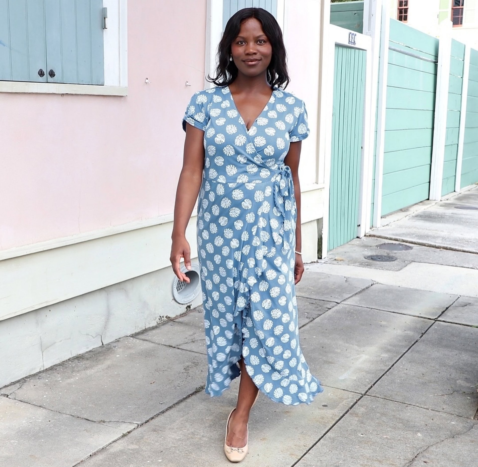 A woman wearing a blue, hand-block printed wrap dress by Passion Lilie walks along a pastel-colored New Orleans street. Passion Lilie, rated “Disrupting” by Sustainable Jungle, is one of our top sustainable fashion brands known for ethical production and timeless style.