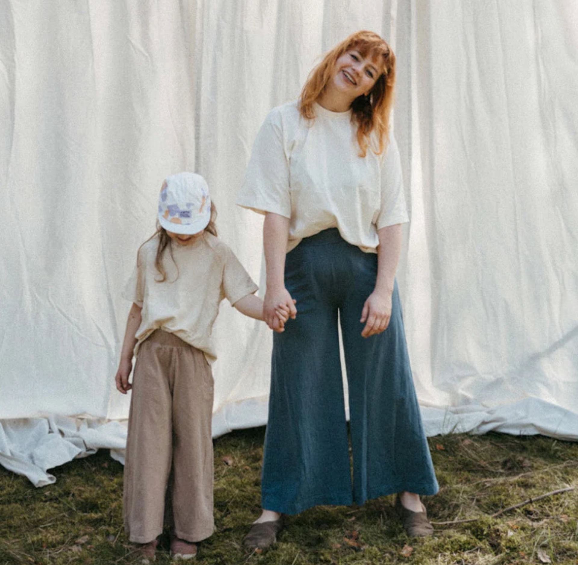 A mother and daughter wearing Orbasics organic cotton outfits in neutral tones stand outdoors holding hands. This image is featured in the Sustainable Jungle Brand Directory listing for Orbasics.