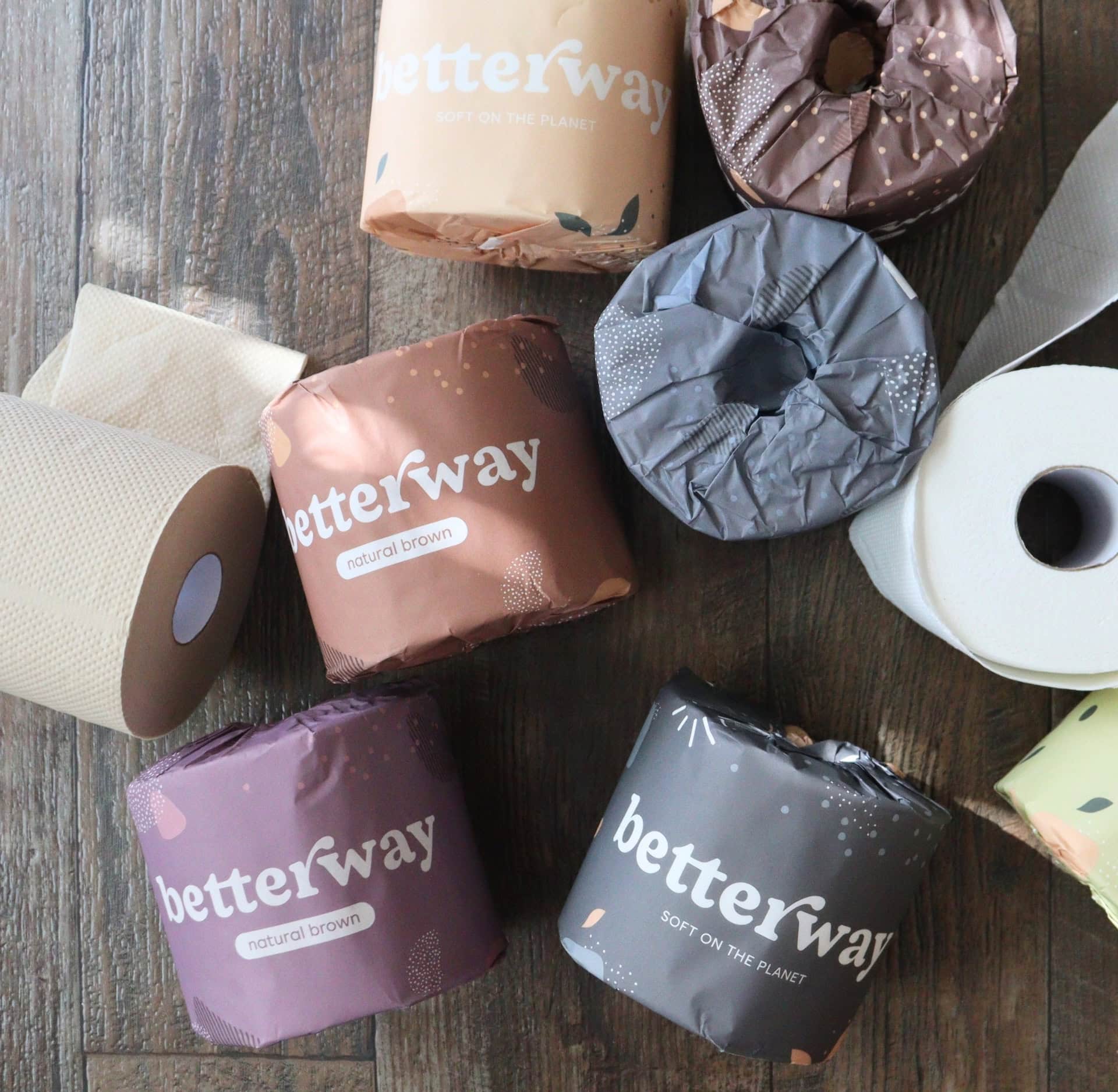 Collection of Betterway bamboo toilet paper rolls in assorted colored wrappers and unwrapped rolls displayed on a wooden floor, tested by Sustainable Jungle for the Betterway Brand Rating.